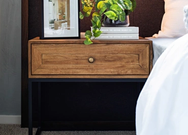 A black metal-framed bedside wooden table with a single drawer is topped with a photo frame & potted plant