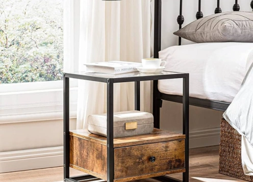 A black metal frame bedside table with a rustic wooden drawer & glass top, decorated with a small box & book.