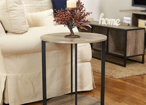 A round wooden side table with metal legs beside a white sofa, decorated with a candle and flowers.