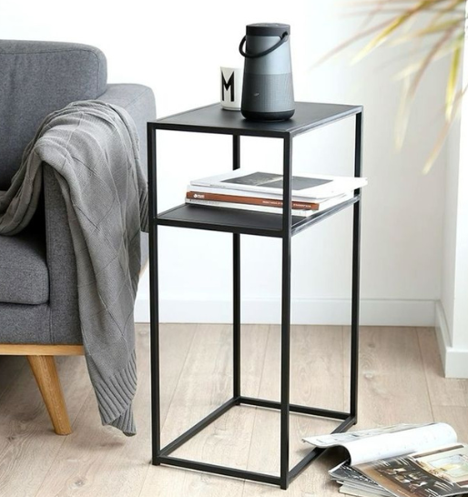 A modern black side table with a speaker on top next to a grey color sofa and a draped throw.